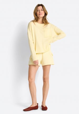 Woman stands with one hand on hip, wearing light yellow long-sleeve top with pocket, matching shorts, and brown flat shoes.