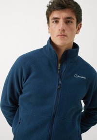 Marine fleecejakke med høy krage, glidelåsfron og sidelommer. Har Berghaus-logoen på brystet. Myk tekstur.