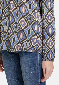 Langärmlige Bluse mit geometrischem Muster in Blau, Beige und Bordeauxrot. Kombiniert mit Jeans aus Denim, die mit kleinen dekorativen Perlen verziert sind.