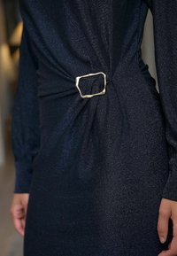 Must mustavärvi särav kleit, millel on vöökohtades keeratud disain, rõhutades metallilist ebaühtlase kujuga pandlit kui silmapaistvat aksessuaar.