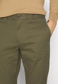 Zielone oliwkowe spodnie bawełniane o gładkiej fakturze, z zapięciem na guziki z przodu i kieszeniami bocznymi, część swobodnego stroju.