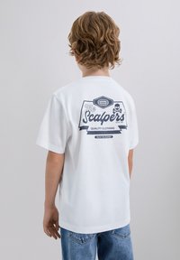 Valkoinen puuvillapaita, jossa on graafinen painatus selässä, jossa on "The Scalpers", kallo ja nauha. Slim fit -malli lyhyillä hihoilla.