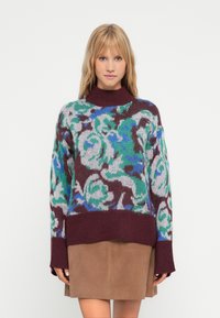 Maglione lavorato a maglia con motivo floreale in blu, verde e bordeaux. Collo alto, maniche lunghe e vestibilità rilassata, abbinato a una gonna in suede marrone.