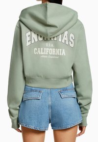 Persona con sudadera corta color verde salvia con el texto "ENCINITAS CALIFORNIA" y pantalones cortos de mezclilla azul claro, vista desde la parte de atrás.