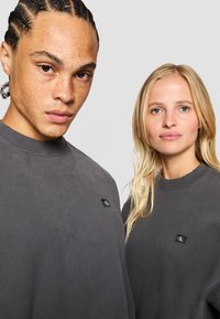 Šedé bavlnené mikiny s rebrovanými výstrihmi, s malou čiernou logovou nášivkou na hrudi. Čistý, minimalistický dizajn, unisex strih.
