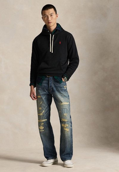 Young man standing wearing a black hoodie over green plaid shirt, distressed blue jeans, white sneakers, and a wristwatch against a neutral background.