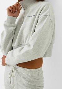 Felpa grigia chiara cropped con colletto alto, chiusura con zip e polsini a coste. Presenta il logo "Twist angel" in blu scuro. Tessuto morbido.