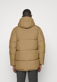 En pustjakke i en lysbrun nuance med en quiltet design, der har hætte og lange ærmer, lavet af et glat, tekstureret stof.