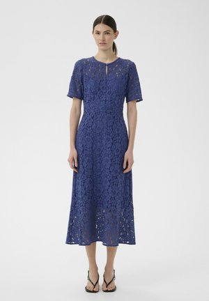 Naine kannab põlvpikkust sinist pitskleiti lühikeste varrukatega ja väikese võlvaugukaelusega, millel on mustad avatud varbaga kontsakingad.