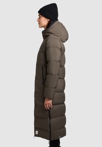 Vrouw in een lange, gevoerde bruine jas met capuchon en een zwarte beanie, staande in profiel tegen een effen achtergrond.