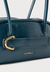 Τσάντα χειρός από πετρόλ δέρμα με διπλές λαβές, χρυσή πόρπη και χρυσό λογότυπο "Coccinelle" σε λείο φόντο.