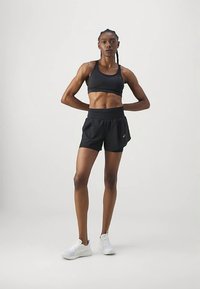 Mulher atlética em pé com as mãos atrás das costas, vestindo um top desportivo preto, calções pretos e sapatilhas brancas, sobre um fundo claro e simples.