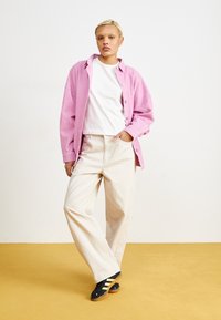 Rosa kordfläds skjorta, vit T-shirt, krämfärgade vida byxor och svarta sneakers med gula ränder. Bakgrunden är gul.