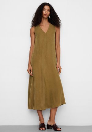 Kobieta o kręconych włosach, ubrana w oliwkową, bezrękawnikową sukienkę midi i ciemnobrązowe mule na obcasie, stojąca na białym tle.