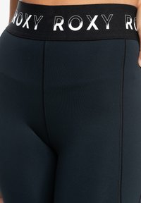 Fascetta in bianco e nero con logo Roxy, leggings scuri aderenti, texture liscia, dettaglio della cucitura laterale e materiale elasticizzato progettato per il comfort.