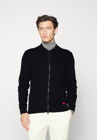Black zip-up sweater with a ribbed collar and cuffs, featuring a subtle texture and a small red brand tag on the lower side.