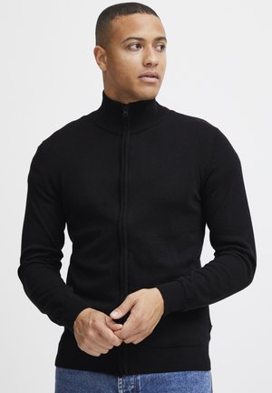 Svart zip-up cardigan med høy krage, ribbede mansjetter og hem. Stoffet ser glatt ut med en tettsittende design. Brukt over denim.