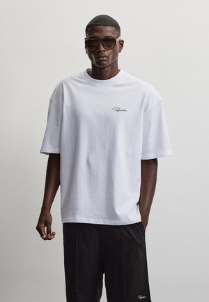 Uomo che indossa una maglietta bianca oversize e pantaloni neri con occhiali da sole, in piedi davanti a uno sfondo grigio chiaro uniforme.