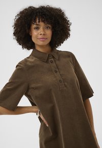 Brązowa sukienka koszulowa z sztruksu, z krótkimi rękawami, listwą guzikową i kołnierzykiem. Materiał ma teksturowany wygląd, co dodaje wizualnego zainteresowania.