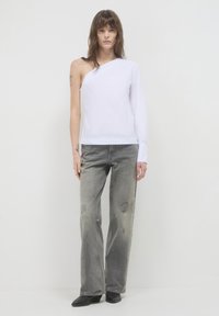 Frau trägt eine asymmetrische weiße Bluse mit einem langen Ärmel und grauen, abgetragenen Jeans, die vor einem einfarbigen, hellen Hintergrund steht.