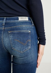 Tummansiniset farkut keskitason vyötäröllä, joissa on takataskussa erottuva kaareva ommel ja vyötäröllä merkintälappu.