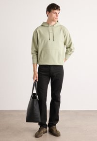 Sudadera verde con capucha y bolsillo frontal, jeans negros y zapatillas marrones. Bolsa de mano negra con dos asas en la mano, de pie en el interior.