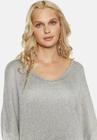 Sidabrinis V formos kaklo megztinis su laisvu pjūviu ir tekstūruotu mezgimu. Yra švelnus metalinis blizgesys, derinamas su spalvingomis kabinančiomis auskarais.