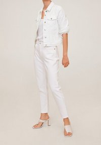Chaqueta de mezclilla blanca con cierres de botones y bolsillos en el pecho, combinada con jeans skinny blancos y sandalias de tacón beige, sobre un fondo neutral.