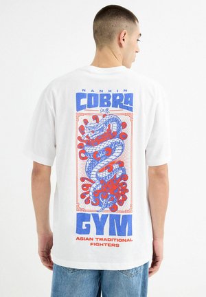 Camiseta blanca con un gran gráfico azul y rojo de una serpiente estilizada y el texto "GIMNASIO NANKIN COBRA LUCHADORES TRADICIONALES ASIÁTICOS" en la parte posterior.
