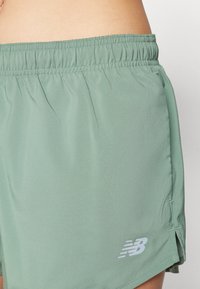 Vihreät urheilushortsit, joissa on joustava vyötärö ja sivurei'ät. Alhaalla vasemmalla on hienovarainen valkoinen "New Balance" -logo.