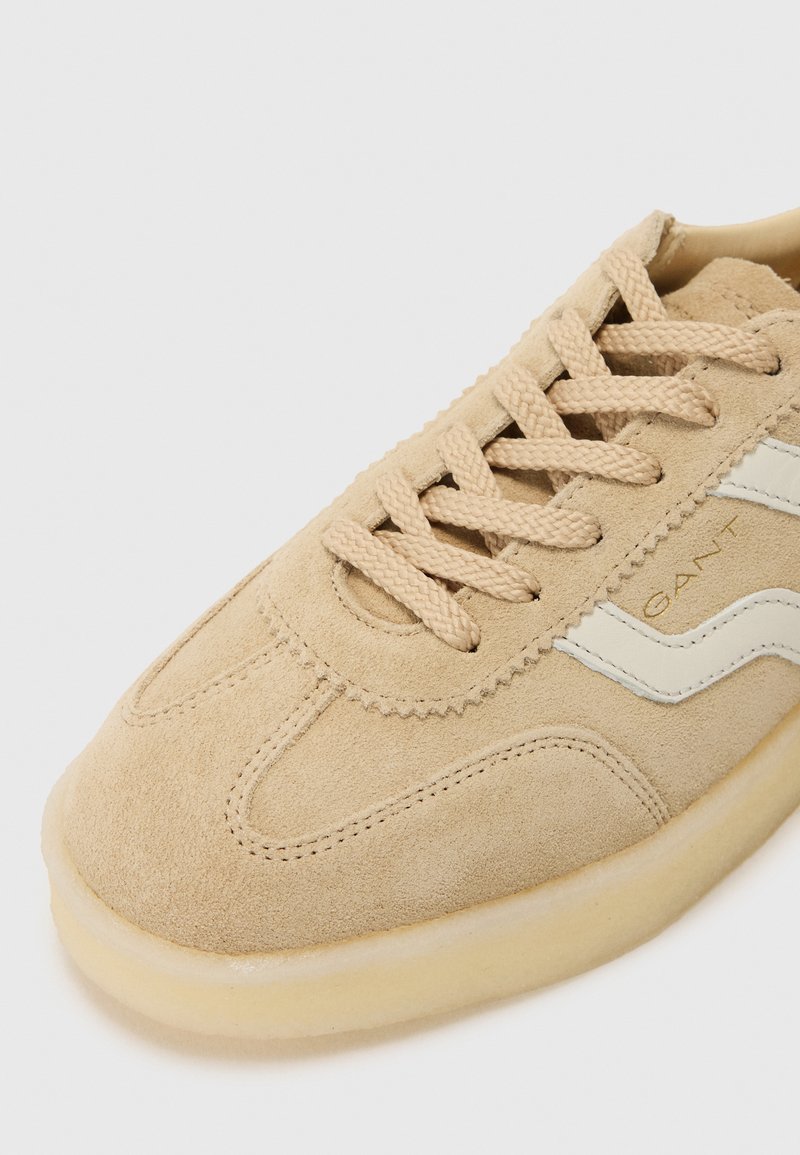 Beige Wildleder-Sneaker mit beigen Schnürsenkeln, weißen Seitenstreifen und einer strukturierten Gummisohle. Verfügt über Stichdetails entlang des Obermaterials.