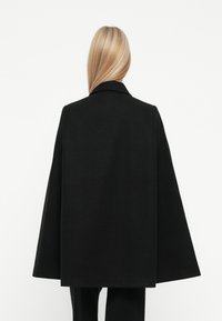 Cappotto a mantello in misto lana nero con una silhouette ampia, colletto a punta e tessuto testurizzato. Cuciture visibili lungo i bordi.