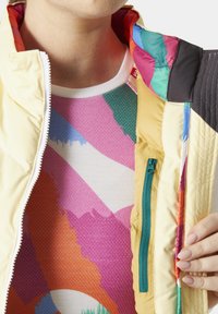 Izolovaná žlutá bunda s vícetrobným interiérem, zipovou kapsou a geometricky vzorovanou košilí pod ní s živými barvami.