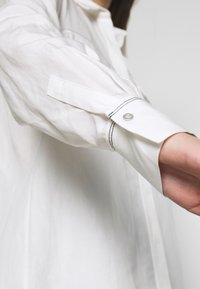 White collared shirt with long sleeves, featuring contrasting dark stitching along cuffs and a snap button detail at the sleeve openings.