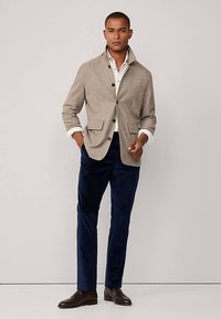 Szara marynarka z mieszanki wełny z dwoma przednimi kieszeniami, guziki, kremowa koszula pod spodem, zestawiona z granatowymi welurowymi spodniami i brązowymi butami.