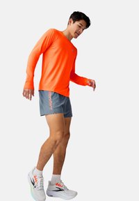 Orange långärmad tröja och grå shorts med "BROOKS" tryckt. Modellen bär grå löparskor med vit bas och färgglada detaljer.