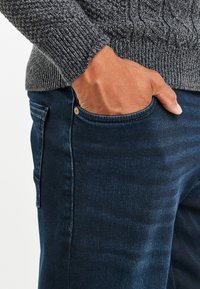 Tummat farkut ovat klassisella leikkauksella ja sileällä pinnalla, joissa on etutasku, jossa on kuparinvärinen nappi. Neulepaidassa on harmaa neulontakuvio.
