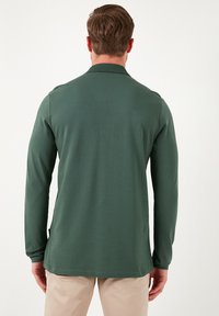 Camisa de polo verde de manga larga, textura suave, cuello de canalé, dobladillo recto, con una etiqueta en el lado izquierdo, combinada con pantalones beige claros.