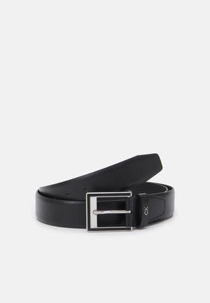 Ceinture en cuir noire avec une boucle rectangulaire argentée et le logo "ck" près de la boucle, roulée et placée sur un fond blanc.