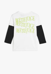 Hvid langærmet skjorte med sorte ærmer; har gentaget grønt tekst "BEETLEJUICE" i forskellige størrelser på ryggen. Blødt bomuldsstof.