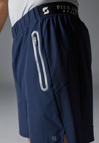 Marineblå atletikshorts med elastisk talje, sidelomme med lynlås og kontrasterende grå detaljer. Letvægtsstof med en glat tekstur.