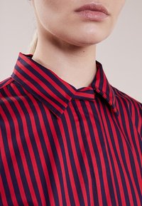 Blusa verticale a righe rosse e blu navy con colletto classico, tessuto morbido e sottile dettaglio con bottoni sulla patta.