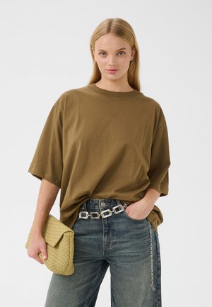 Frau mit langen blonden Haaren, die ein übergroßes olivgrünes T-Shirt trägt, blaue Jeans mit silbernem Gürtel, die eine gewebte beige Clutch hält.