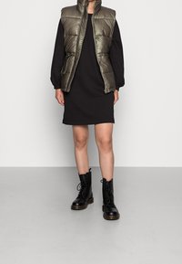 Oliwkowa, wypełniona kamizelka z wysokim kołnierzem na czarnej sukience z długim rękawem. Czarne, lace-up buty wojskowe. Gładkie, błyszczące tekstury, minimalistyczny design.