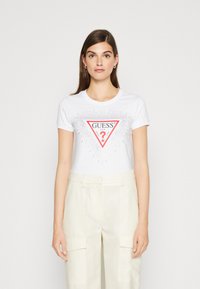 Hvid t-shirt med et rødt trekantet logo med "GUESS" og et spørgsmålstegn, pyntet med små metalliske prikker. Kombineret med lyse cremefarvede bukser.