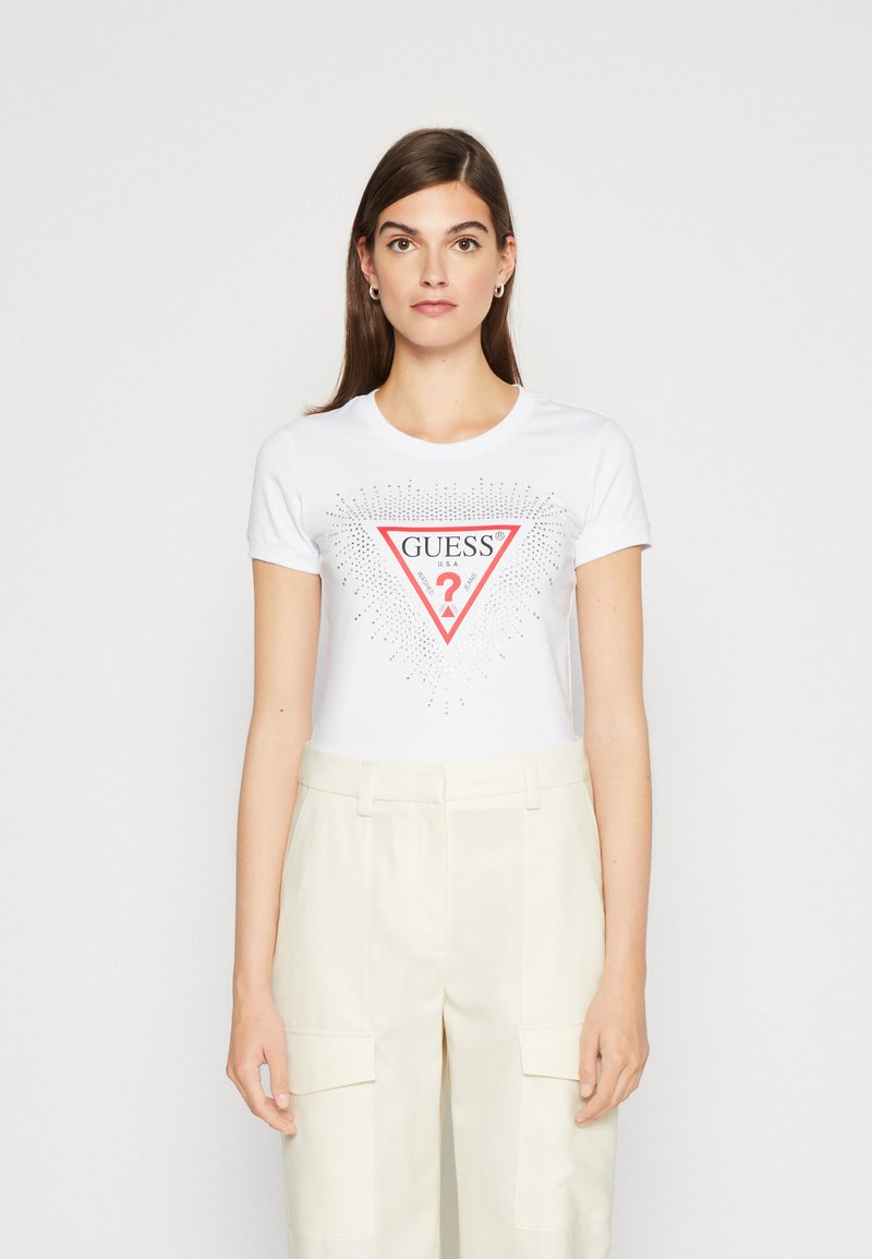 Hvid t-shirt med et rødt trekantet logo med "GUESS" og et spørgsmålstegn, pyntet med små metalliske prikker. Kombineret med lyse cremefarvede bukser.