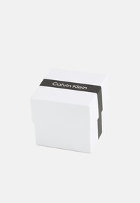 Boîte carrée blanche avec une bande noire portant le texte "Calvin Klein". Texture lisse, design épuré et style minimaliste.