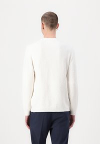 White textured sweater with long sleeves, round neckline, and ribbed cuffs, paired with dark trousers, shown from the back.
