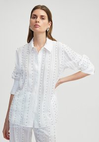 Witte blouse met verticale streepjespatroon, versierd met kleine zilverkleurige studs, met een kraag en losse mouwen.