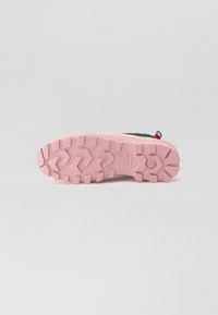 Zapatilla multicolor que presenta una parte superior verde de tela texturizada, suela de goma rosa con un patrón en zigzag y una pestaña para facilitar el calzado.